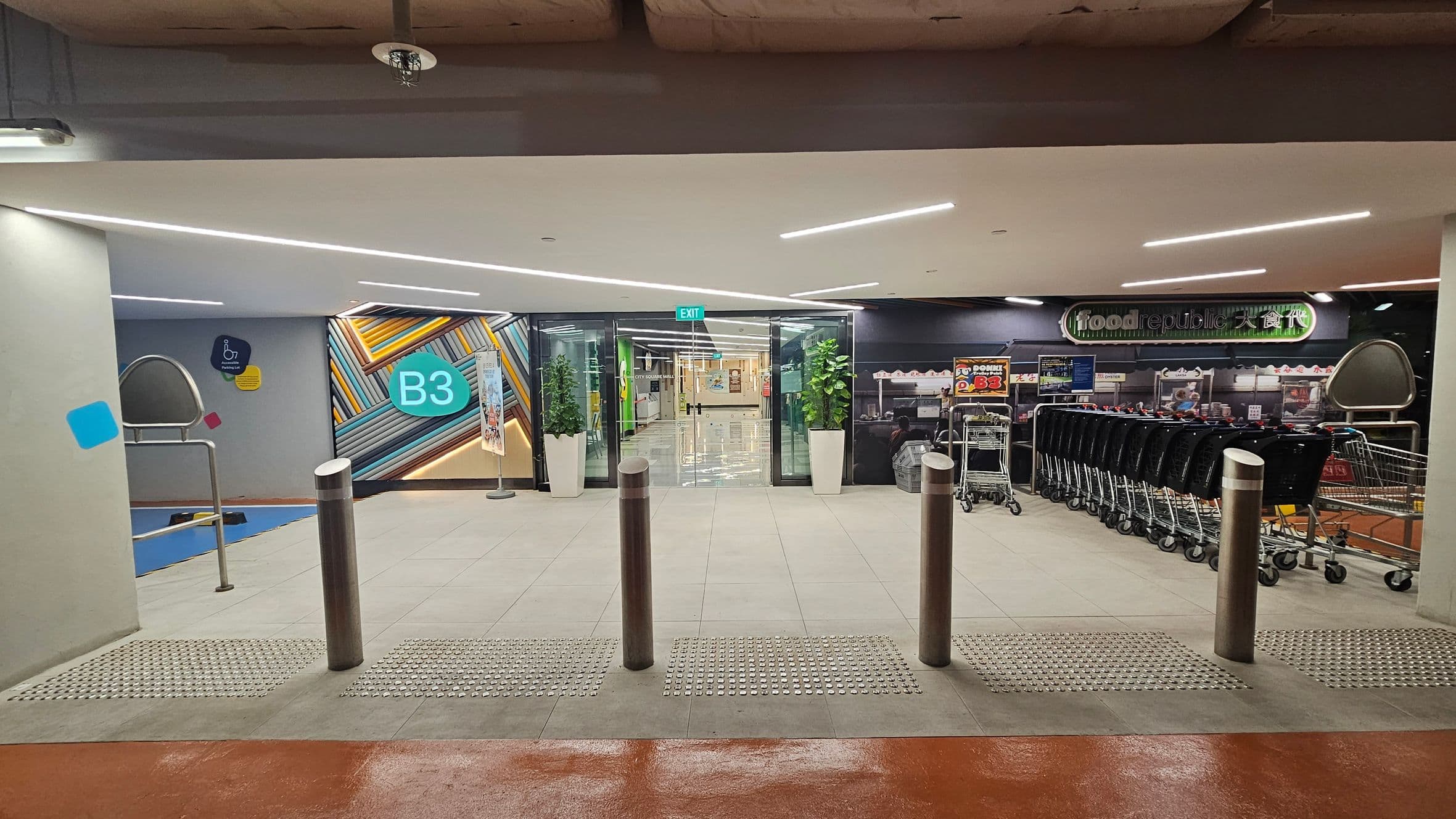 On the left, a wall with a colourful geometric design and a sign marked "B3". In the centre, glass doors with an "EXIT" sign above, flanked by potted plants. On the right, a row of shopping trolleys and a sign for "food republic". The floor is tiled with tactile paving near the entrance, and the ceiling has modern strip lighting.