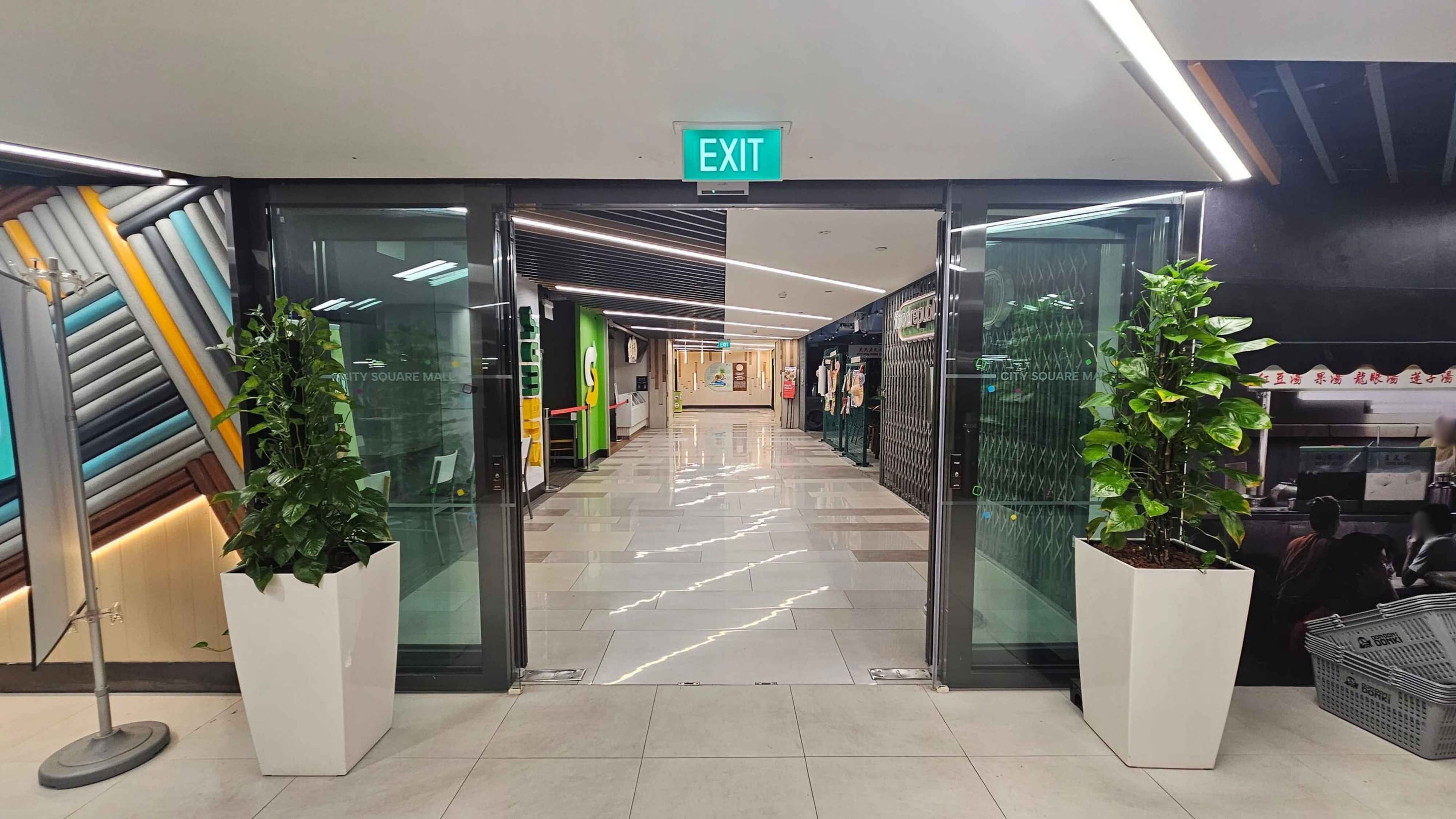 Glass doors with an "EXIT" sign above them, flanked by tall green plants in white pots. Beyond the doors, a corridor with a shiny tiled floor leads to various shops. On the left, colourful diagonal wall patterns, and on the right, a dark wall with a partially visible food stall. The ceiling features linear lighting.