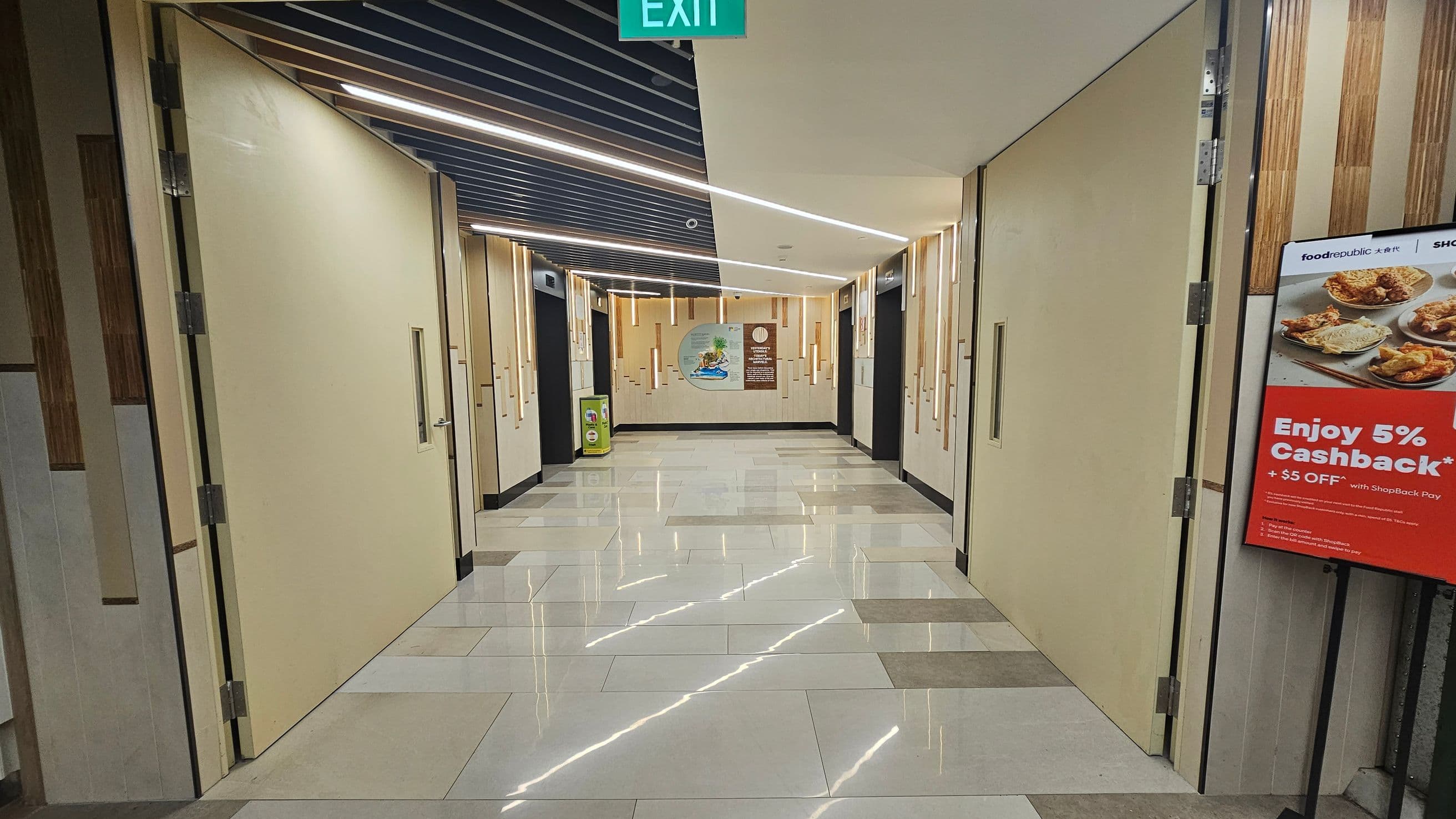 A corridor with beige walls and large doors on both sides. The floor is tiled with a glossy finish, reflecting the ceiling lights. The ceiling has a modern design with linear lights and dark panels. On the right, there's a poster advertising a 5% cashback offer with images of food. At the end of the corridor, there's a mural and a small green bin. An exit sign is visible at the top.