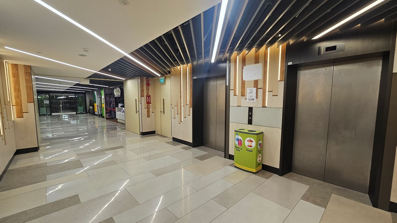 A modern indoor hallway with a polished tiled floor and a ceiling featuring linear lights. On the right, there are two lift doors with a green recycling bin nearby. The left side shows a corridor leading to glass doors, with a fire extinguisher mounted on the wall. The walls have a light wood and beige design, and the ceiling has black slats with integrated lighting.