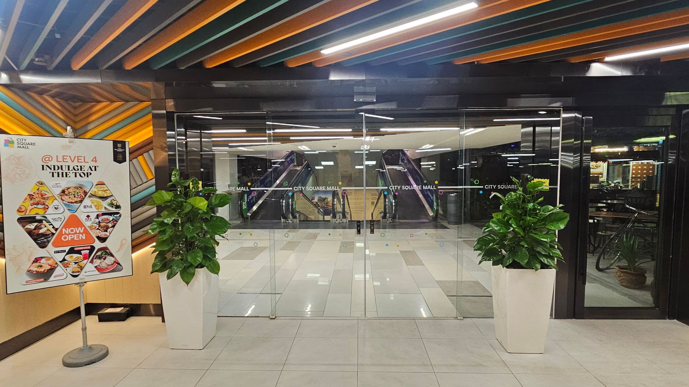 Glass entrance doors to City Square Mall with escalators visible inside. On the left, a colourful sign advertises dining options on Level 4, featuring various dishes and a "Now Open" message. Two large potted plants flank the entrance. The ceiling has a pattern of orange, green, and black slats, and the floor is tiled in light colours.