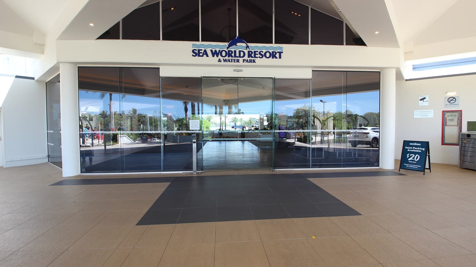 The main drop off area for the Sea World Resort.  The entrance is a series of glass panels and double sliding door.   Above the doors reads the sign "Sea World Resort and Water Park".  The large tiled floor outside is brown with a contrasting colour of tiles changing to black closest to the sliding doors.