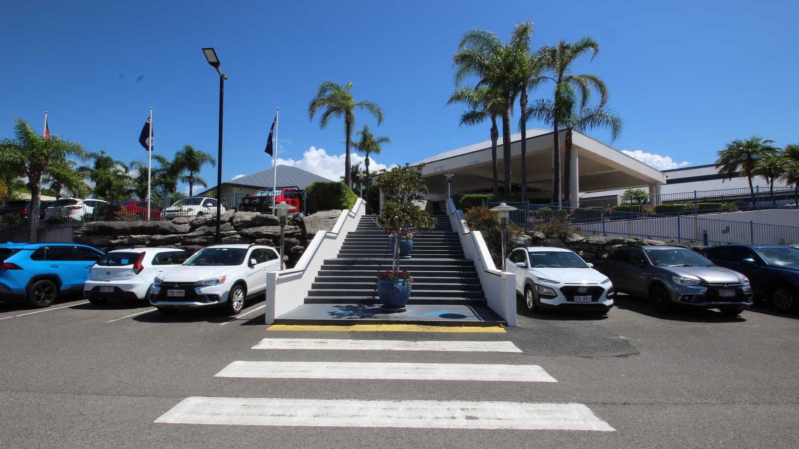 Situated in the main carpark of the Sea World Resort looking up a large flight of stairs toward the entrance of the resort.  The stairs have three tiers moving in same direction and an accessible ramp is evident on the right hand side of the image.  Either side of the stairs are numerous parked cars.