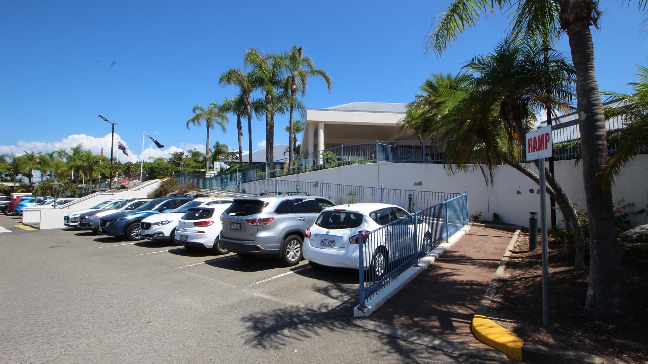 Situated in the main carpark of the Sea World Resort looking up an accessible ramp toward the entrance of the resort.  Toward the left of the image is an evident set of stairs with three tiers moving in same direction toward the entrance of the resort.   Numerous parked cars dominate the foreground of the image.