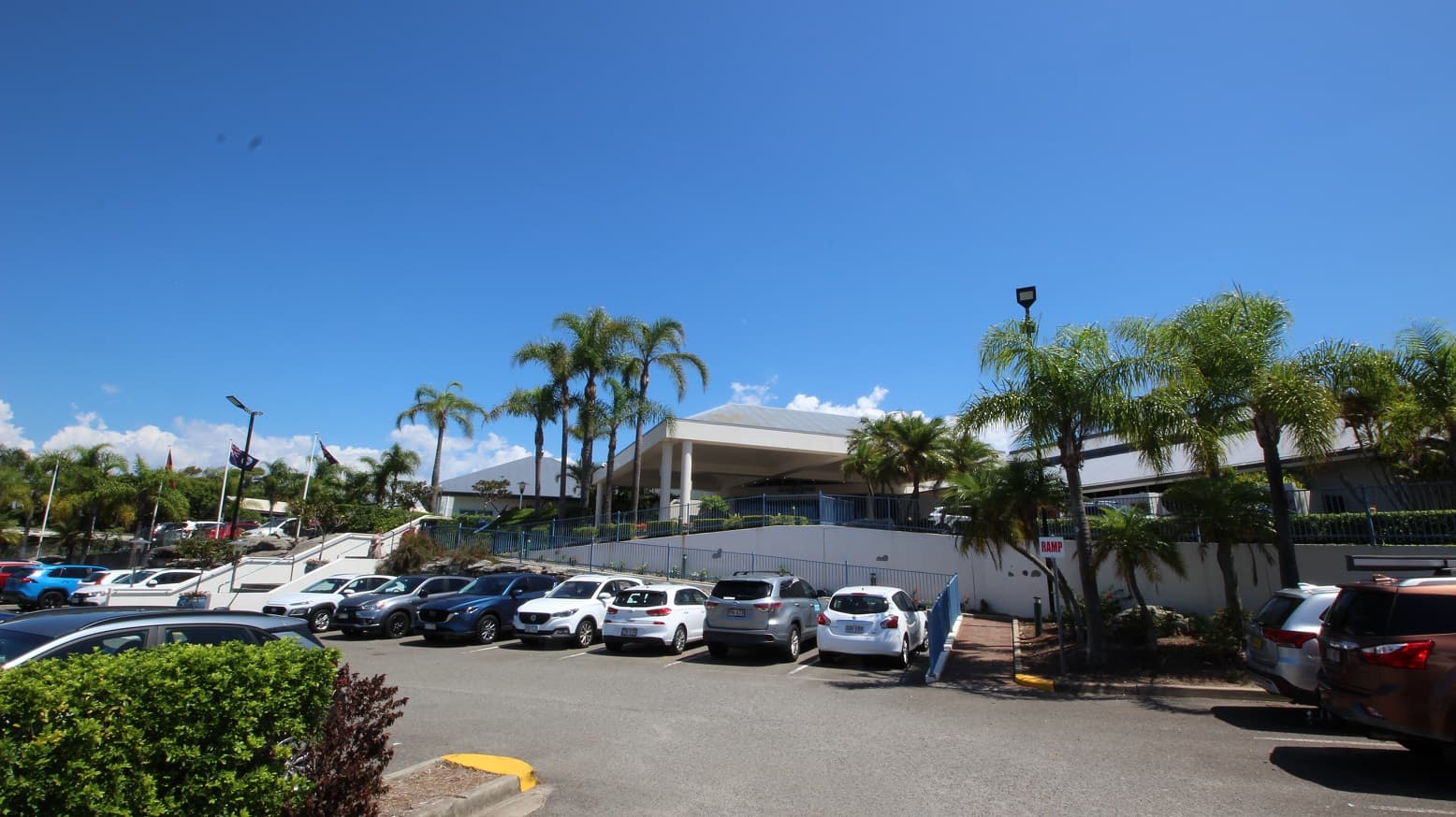 Situated in the main carpark of the Sea World Resort looking up an accessible ramp toward the entrance of the resort.  Toward the left of the image is an evident set of stairs with three tiers moving in same direction toward the entrance of the resort.   Numerous parked cars dominate the foreground of the image.