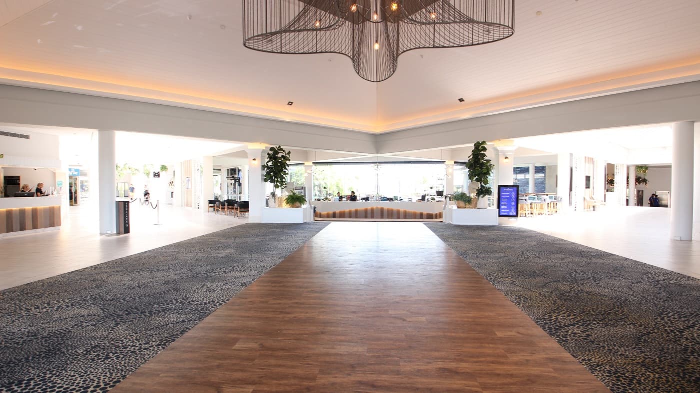 The reception area of the Sea World Resort is very spacious and with high ceilings.  Patterned carpets are the immediate floor covering with white tiled areas filling a reception and corridor space.  On the left wall is the word Reception and a dolphin design above the text of which is back lit to help set a calm mood for the space.  The Reception counter close by is staffed with service personnel.