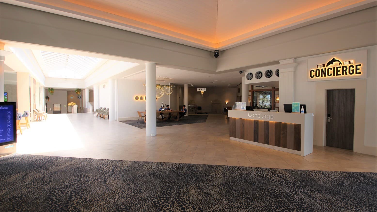 The concierge area of the Sea World Resort is very spacious and with high ceilings.  Patterned carpets are the immediate floor covering with white tiled areas filling a reception and corridor space.  On the right wall is the word Concierge and a dolphin design above the text of which is back lit to help set a calm mood for the space.  Further past the concierge desk is a small sitting area with three people talking casually.