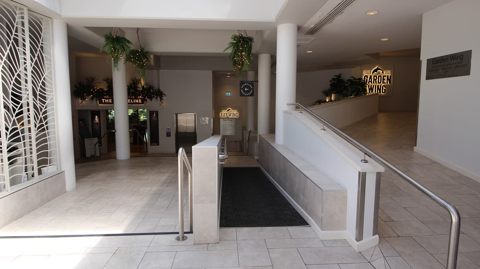 A series of accessible ramps and stairs dominate the image to showcase options to move downwards to the Baywing area or upwards toward the Garden Wing. A small set of three steps are toward the left and a ramp in middle both going down.  Toward the right is an up ramp moving toward a sign reading Garden wing.   The floor is tiles except for the carpeted ramp.
Handrails are evident on all stairs and ramps.