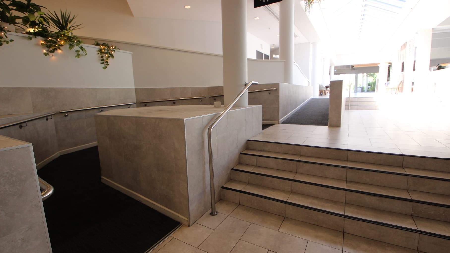 A series of accessible ramps and stairs dominate the image to showcase options to move upwards toward the entrance of Sea World Resort. A small set of three steps are toward the right and a ramp in middle and curves up and around a large white concrete pillar.   All floor coverings are cream coloured tiles and the upramp is dark blue carpet.
Handrails are evident on all stairs and ramps.