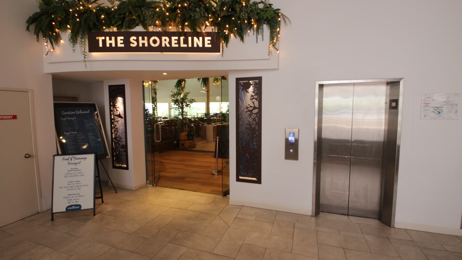 Entrance into the Shoreline restaurant with an open set of double glass doors.  Toward the right is a stainless steel elevator doors.  Ouside of the restaurant the floor coverings are a cream tile and directly inside is a dark brown timber flooring.  Above the double door entrance are the the 
The Shoreline signage that is backlit.  A restaurant menu and specials board are situated to the left of the entrance doors.