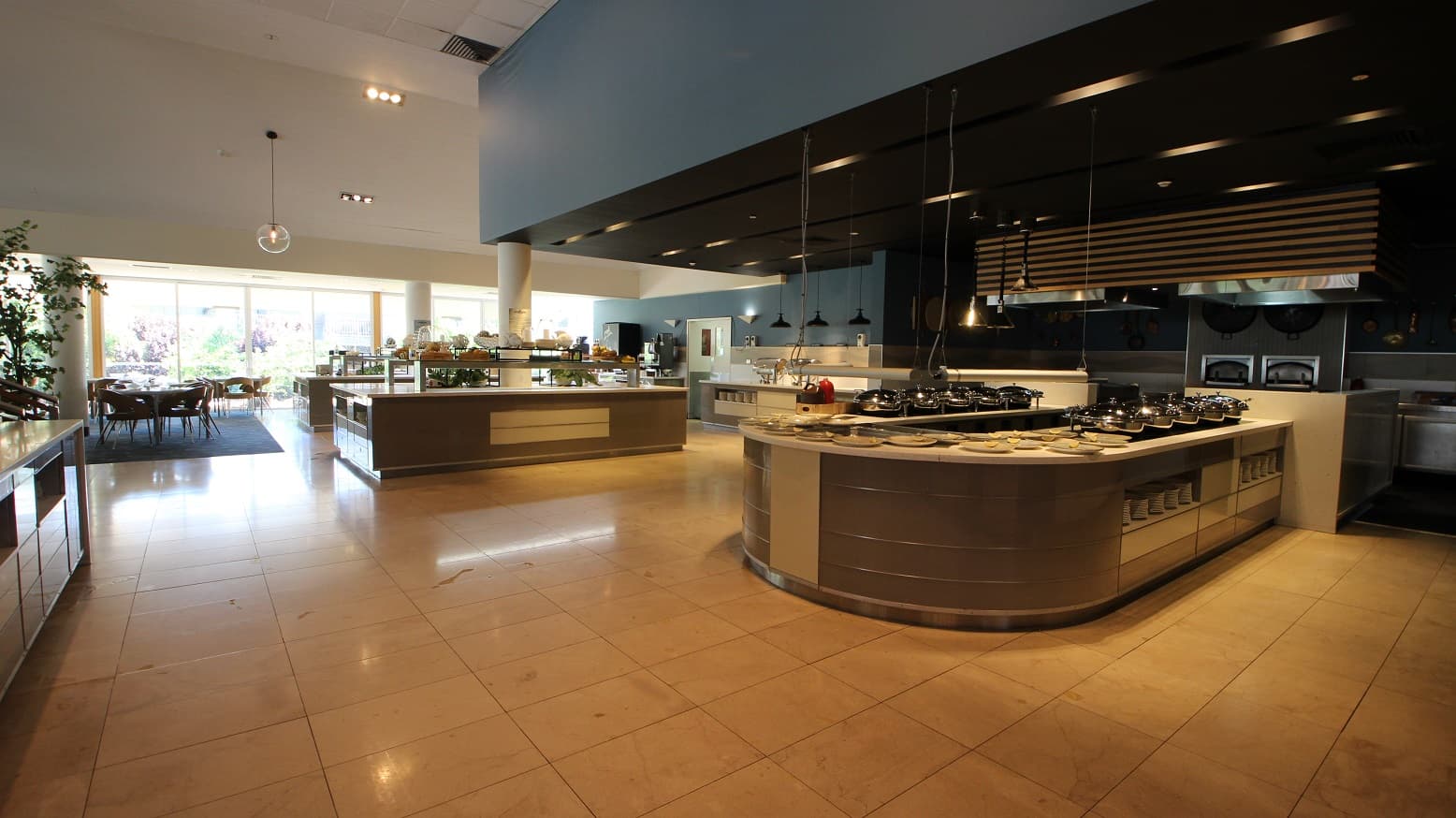 The buffet and servery areas within The Shoreline restaurant.  One U shaped servery bench contains 10 hot bain maree dishes and multiple plates.  A rectangular servery bench toward the left contains various fruit baskets.  The tiled flooring area is a cream/light brown colour.