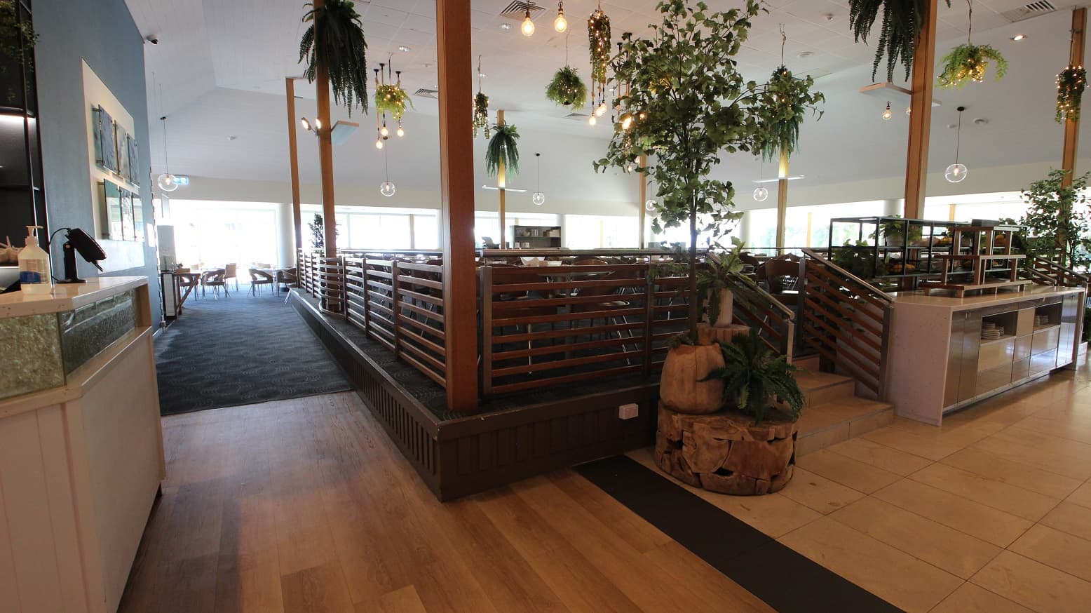 Turning left from the entrance to The Shoreline showcases a corridor toward another series of dining tables and chairs.  The step free corridor transitions from a brown timber flooring toe blue patterned carpet almost halfway down the corridor.  Toward the middle and left of the image is the central elevated 3 step platform to a dedicated dining area with up to 20 restaurant tables and chairs.