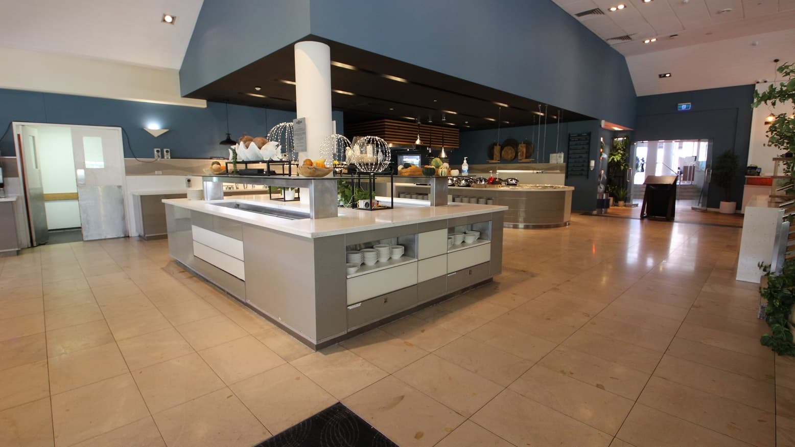A rectangular food servery station within The Shoreline restaurant with white counter tops and grey sides.  Stacks of bowls can be seen below the counter top and various fruit and bread baskets sit atop.  The floor is a large cream tile and high white ceilings.  A section of the servery area has a blue ceiling to separate the servery area from the remainder of the restaurant.