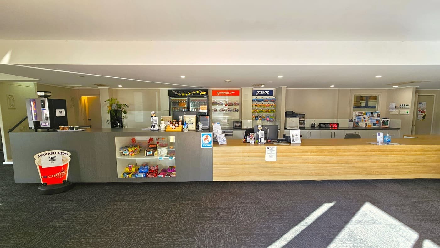 On the left is a cafe presenting snacks and drinks, featuring a coffee maker atop the counter and a fridge for cold drinks at the back. To the right is the reception counter, designed with low timber. Brochures and a computer are neatly placed on the counter, and goggles are arranged in a display rack behind. The low white ceiling is illuminated by LED lights, and the floor is grey. Posters, including an advertising banner in front of the cafe, are affixed to the wall.