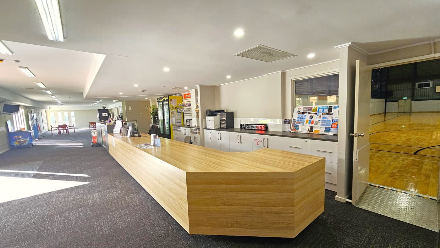 A lengthy reception counter with a timber design. Pamphlets, brochures, and other office equipment are neatly arranged on the grey drawers behind. The floor has grey carpet, an ice cream fridge on the left, and a flatscreen TV mounted to the left wall. Movable tables and chairs are visible. To the right is an entrance to the stadium, featuring polished timber flooring and an aluminum step-free ramp.