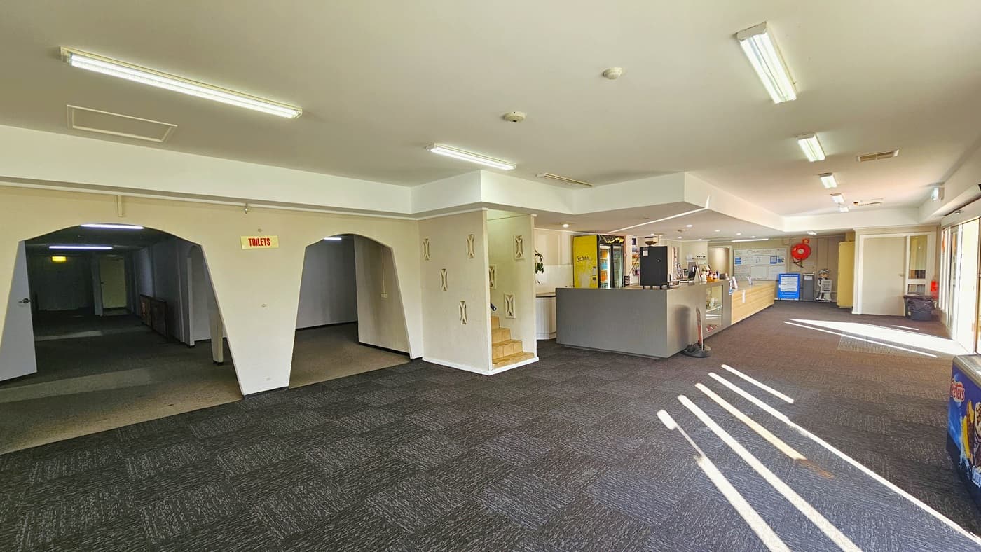 On the left, two dome-shaped entrances with "toilet" signs are visible. The reception and cafe are situated in the middle, featuring a fridge and a coffee vending machine. An ice cream machine is positioned on the right. Further ahead, various signages and posters are displayed. An enclosed space with stairs is located to the left of the cafe. The floor is covered in grey tiles, and the ceiling is entirely white with fluorescent lights.