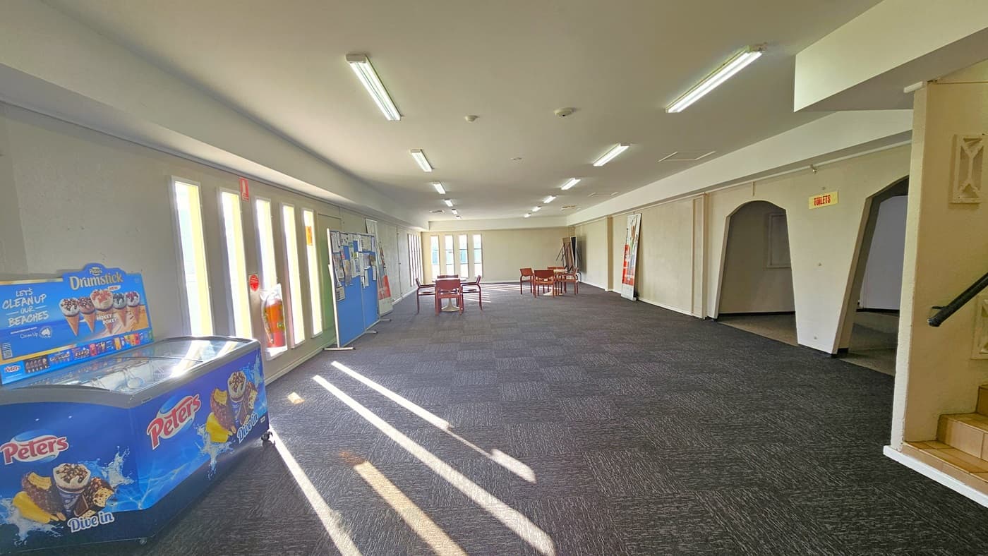 On the right, two dome-shaped entrances with "toilet" signs are visible. An ice cream machine is positioned on the left. An enclosed space with stairs is located to the left. Various signages and posters are displayed on a blue board near the fire extinguisher. Movable red plastic chairs are positioned in the middle. The floor is covered in grey tiles, and the ceiling is entirely white with fluorescent lights.