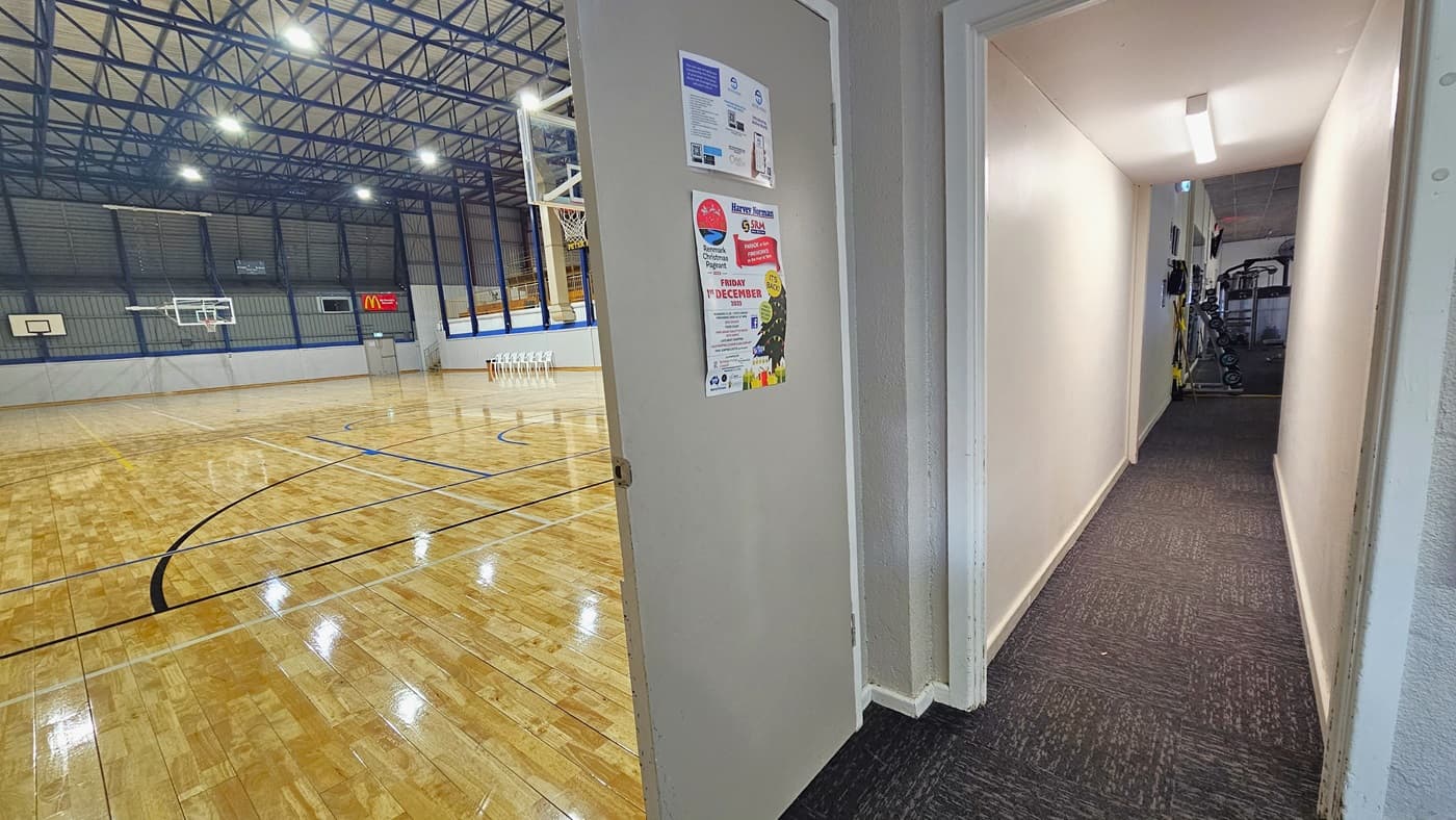 To the left, is the access to the stadium through a grey door. The floor features polished timber with markings, and the ceiling has an industrial look with exposed blue beams. To the right, a narrow corridor leads to the gymnasium. At the corridor's end, exercise machines are visible, with colorful posters affixed to the door.