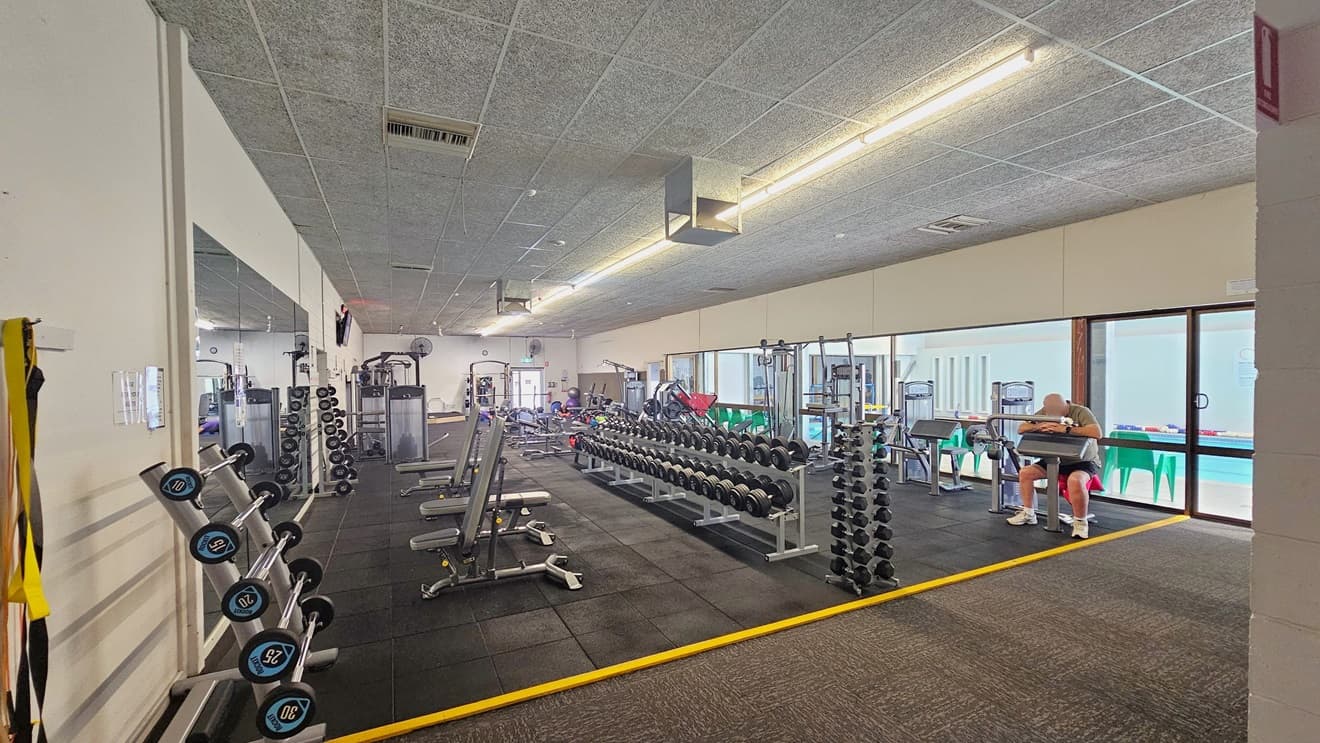 To the left are neatly arranged weight machines, benches with weights, and barbells. On the right, arm curl machines, and further ahead, more exercise machines. A floor-to-ceiling mirror on the left wall, and glass on the right overlooking the pool. The floor transitions from grey carpet to black rubber tiles, with a yellow line. The ceiling has a grey, porous design, and lanyards are hung near the entrance.
