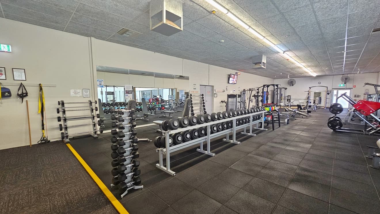 The entire floor is lined with exercise machines. On the left, weightlifting and smith machines are neatly arranged, with a floor-to-ceiling mirror. On the right are rowing machines. The left wall provides space to hang bags, featuring a framed poster and an exit sign. The floor transitions from grey carpet to black rubber tiles separated by a yellow line.