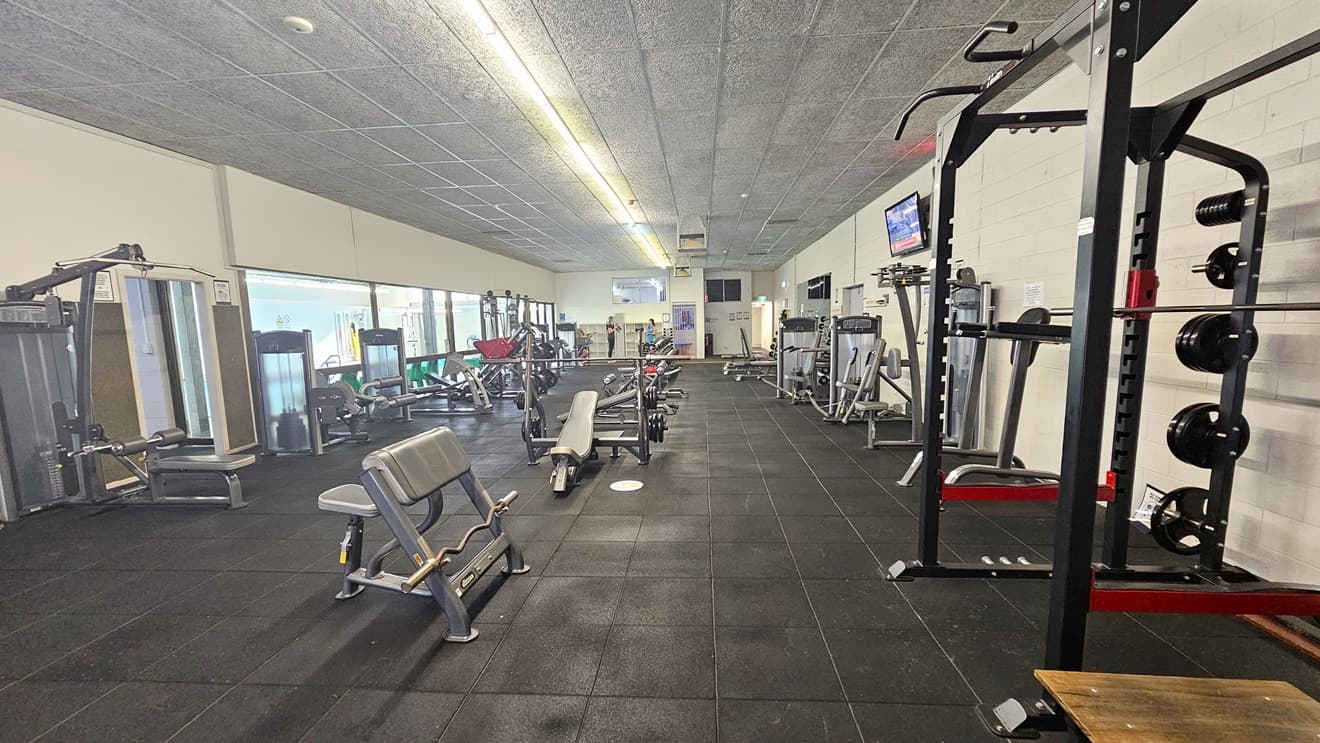 On the left are the overhead press and rowing machines. In the middle, bench press and leg extension machines. On the right, are other machines, including a smith machine. The floor features dark grey rubber tiles, and the ceiling has a porous design in grey. A flat-screen TV is mounted on the right wall. Further ahead is a rack for belongings.