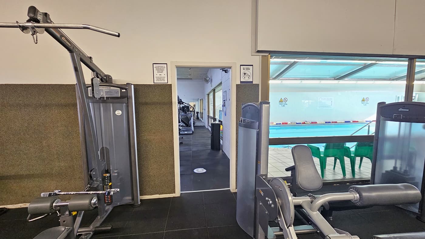 On the left, a lat pulldown machine, and on the right, a leg extension machine. In the middle, an entrance leads to the other side, displaying more machines. A banner is placed on top of the door. The left wall is brown and white, while the right wall is made of glass, overlooking the pool outside.