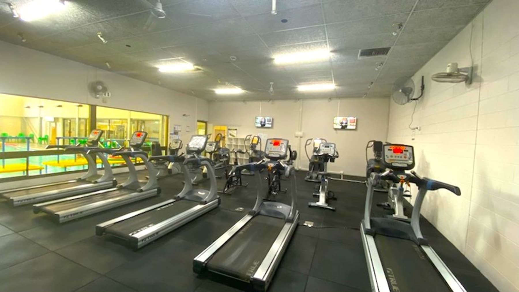 Five treadmills lined up with exercise bikes visible behind these treadmills. To the left are windows and the two other walls are white with fans and TV screens affixed to the walls.