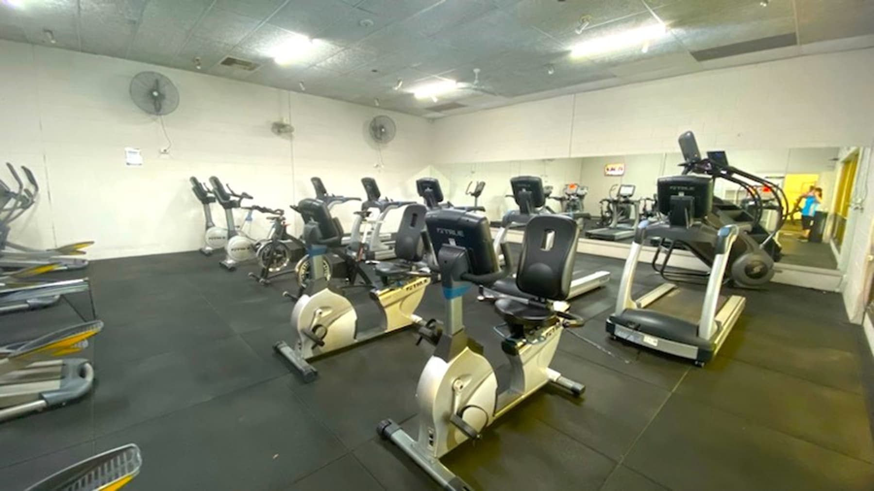 Exercise bikes with treadmills visible behind the bikes. Ceiling fans are affixed to the back wall. The far wall has mirrors affixed to the wall. Fluorescent lights are on the ceiling.