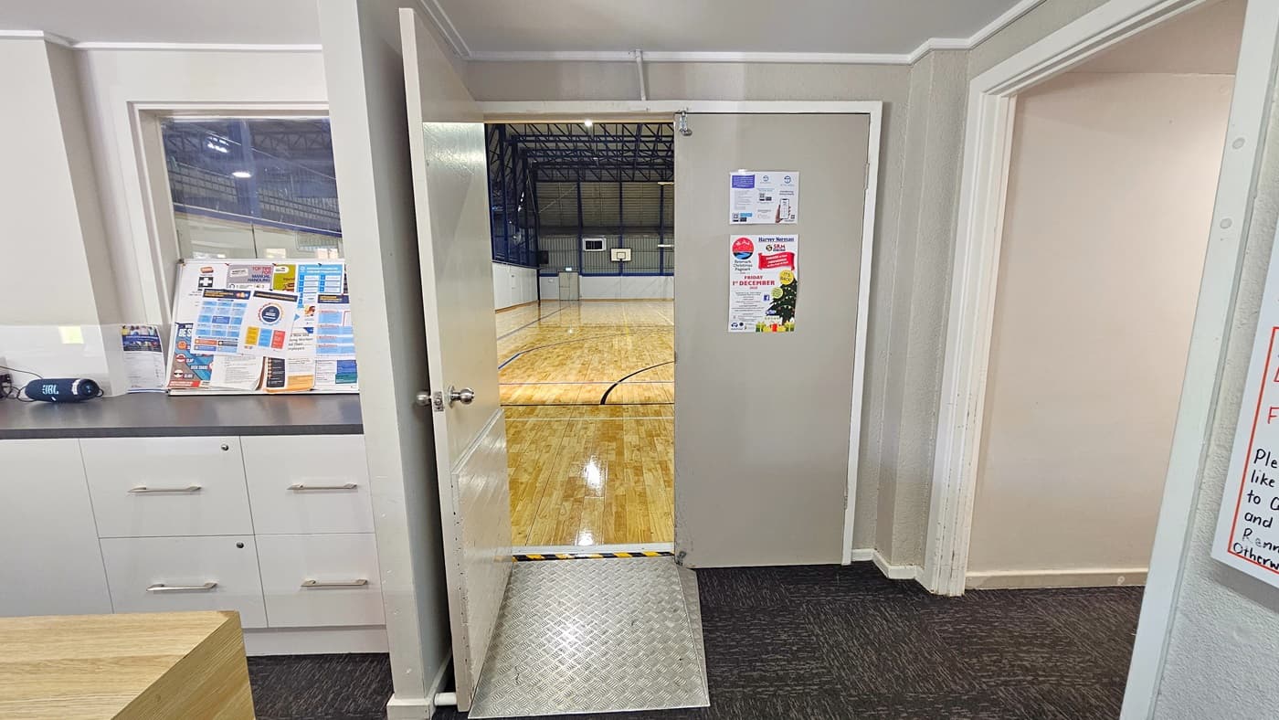 To the left, a section of the reception area is visible. On the right is a corridor leading to the gym. In the middle is a manual door that opens outward, with an aluminum step-free ramp positioned on it. Colorful posters are affixed to the door. Inside, the stadium is visible, featuring timber floors with markings for various ball games, and an industrial ceiling with exposed blue beams.