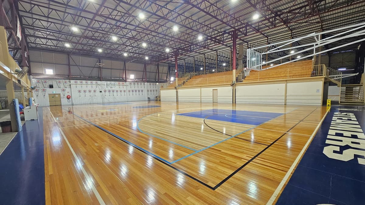 Stadium 1: The first stadium features polished timber floors with markings for various ball games. The left and right sections have blue tiles bearing a team's brand. At the far end, a white corrugated wall displays triangle banners. To the right, the blue tiles lead to the second stadium. Tiered seating in the middle serves as a divider. The ceiling has an industrial look with exposed blue and red metal beams, along with corrugated lining.