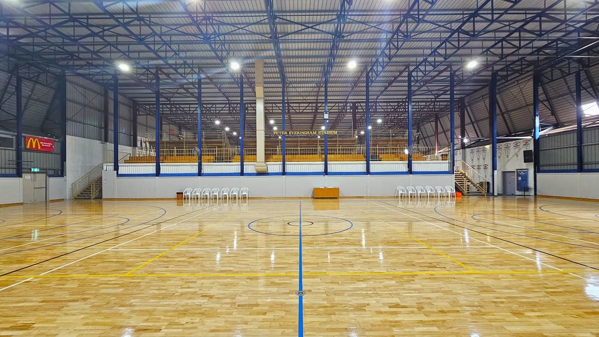 The stadium is equipped with polished timber floors marked for various ball games. In the middle, tiered seating with railings is accessible via wooden stairs with railings. White plastic chairs and a desk are lined up below. The ceiling has an industrial look with exposed blue metal beams, along with corrugated lining. An advertising banner is placed on the upper left, and a digital screen is on the upper right.