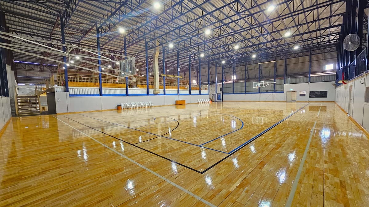 The stadium features polished timber floors marked for various ball games. In the middle, tiered seating with railings is accessible via wooden stairs. White plastic chairs and a desk are lined up below. The ceiling has an industrial look with exposed blue metal beams and corrugated lining. A wall fan is positioned on the middle wall, and basketball boards are placed on both sides.