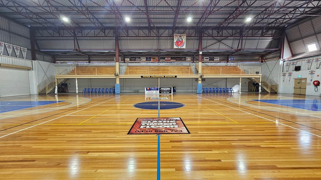 The stadium features polished timber floors marked for various ball games. In the middle, tiered seating with railings is accessible via two wooden stairs. Blue plastic chairs and a desk are lined up below. The ceiling has an industrial look with exposed blue metal beams and corrugated lining. Flyers and banners surround the area. An exit door and a fire hose reel are placed in the right corner.