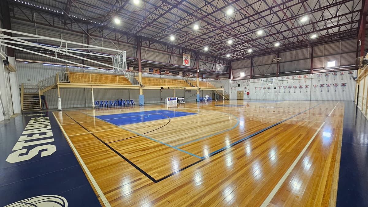The stadium has polished timber floors marked for various ball games. In the middle, tiered seating with railings is accessible via two wooden stairs. Blue plastic chairs and a desk are lined up below. The ceiling has an industrial look with exposed blue metal beams and corrugated lining. Flyers and banners surround the area. An exit door and a fire hose reel are placed in the right corner. Blue tiles with a brand name line the left and right edges of the court.