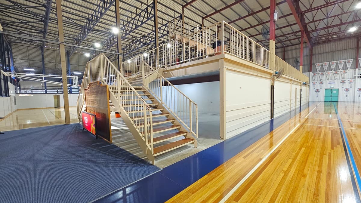 The stadium has polished timber floors marked for various ball games. In the middle, tiered seating with railings is accessible via two wooden stairs. Blue plastic chairs and a desk are lined up below. The ceiling has an industrial look with exposed blue metal beams and corrugated lining. Flyers and banners fill the right wall. An exit door and a fire hose reel are placed in the right corner. To the left is a carpeted area with a large brown board, below which is an advertising banner.