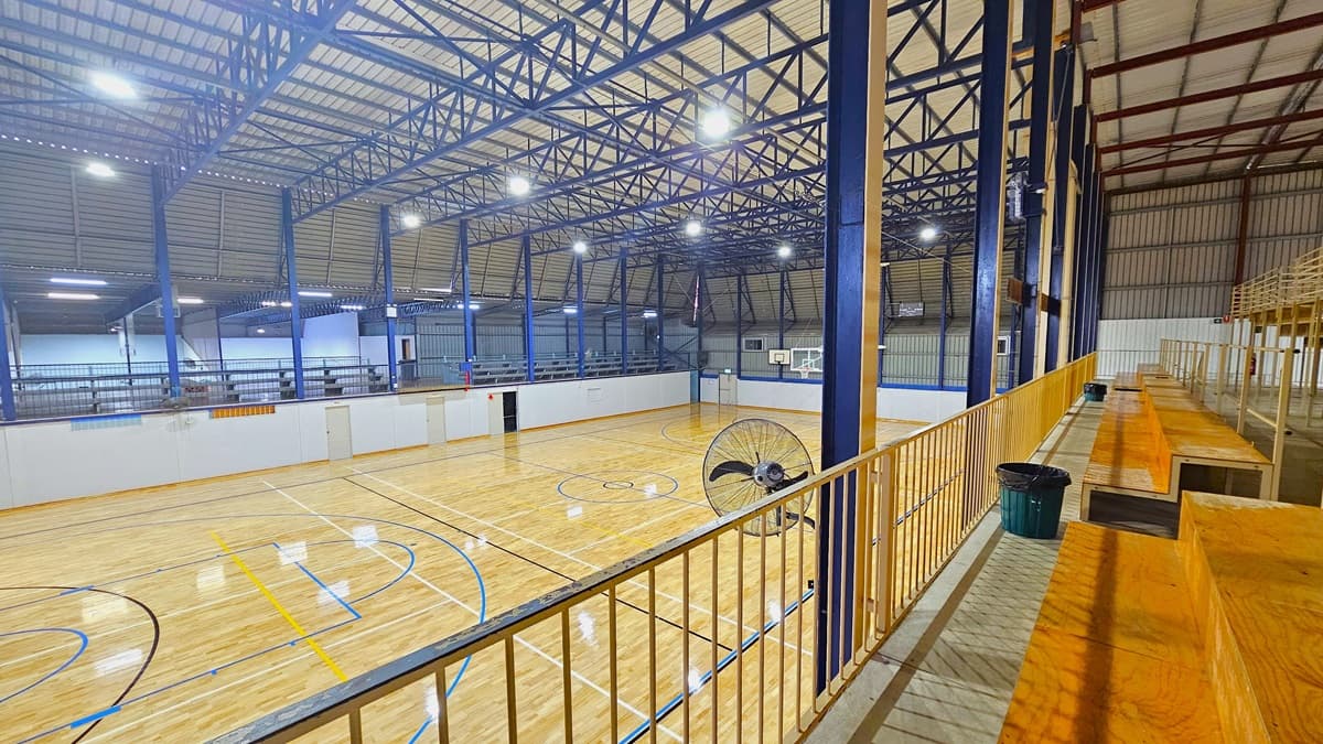 To the right, tiered seating made of timber features steel railings for security. Rubbish bins are placed at the foot of the steps. A wall fan and flat-screen TVs are mounted on the wall. To the left is the court, with a timber floor marked for various ball games. On the other side, there is another tiered seating area. The ceiling has exposed blue and red beams with corrugated lining.