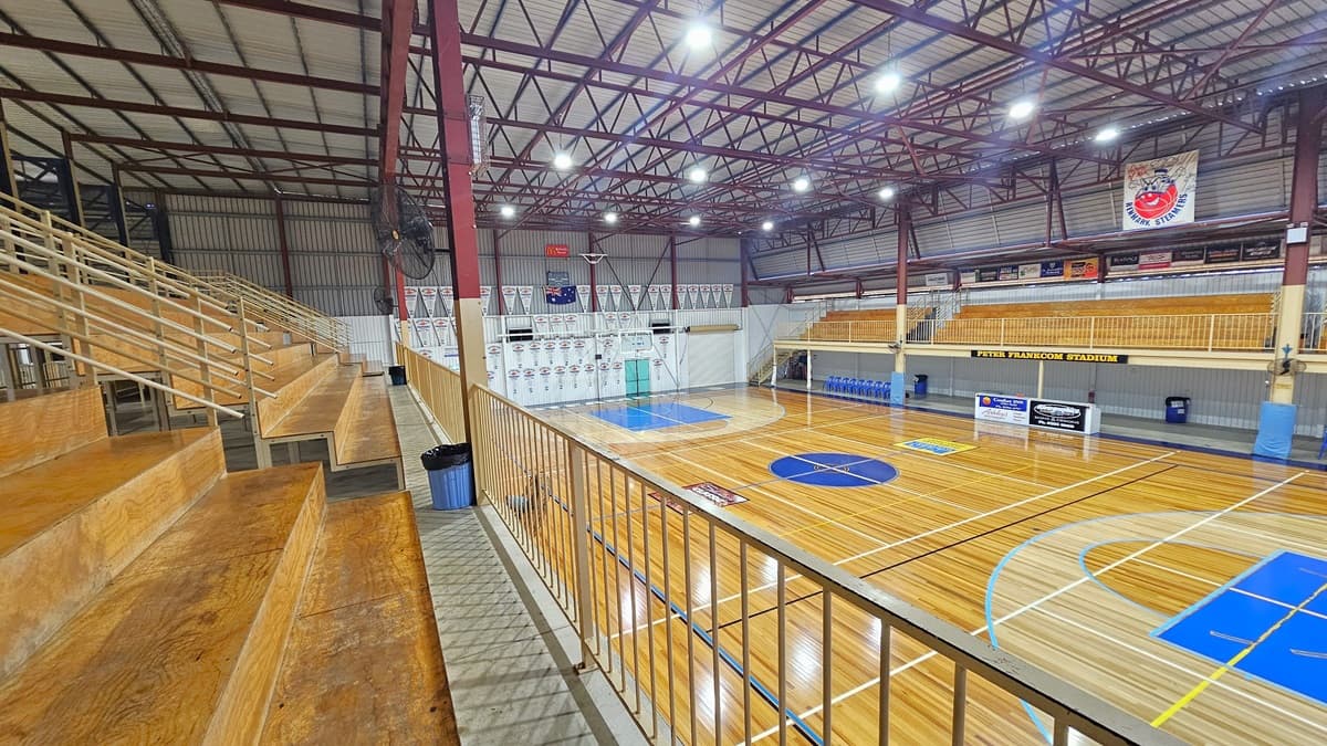 A close view of the tiered seating reveals timber construction with steel railings for security. Rubbish bins are positioned at the foot of the steps. On the wall, a fan and flat-screen TVs are mounted. To the right is the court, featuring a timber floor marked for various ball games. On the opposite side, there is another tiered seating area. The ceiling displays exposed red beams with corrugated lining. Banners and advertising signs are visible.