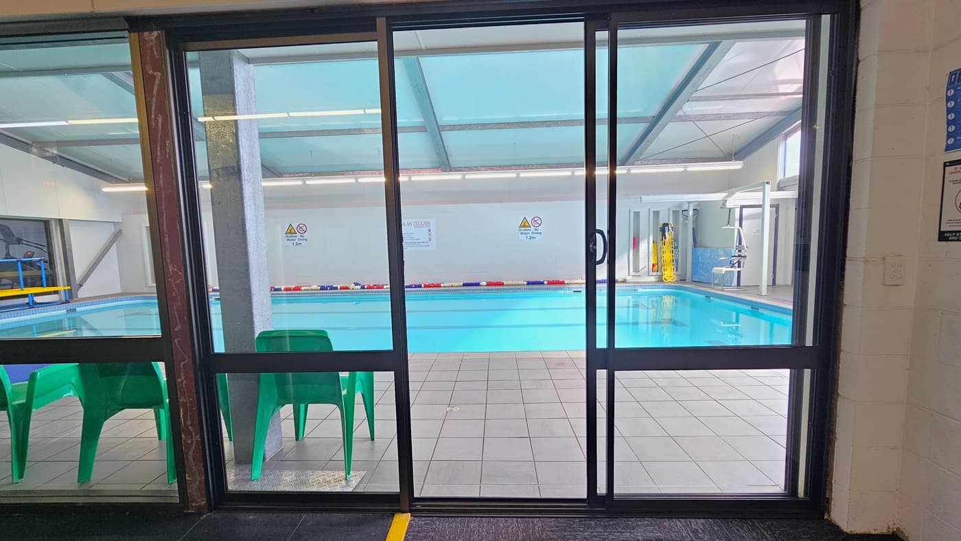 The entrance to the pool is through a glass sliding door. Green plastic chairs are lined up on the left side. The 16m swimming pool features colorful floating ropes. Banners, and safety reminders are affixed to the middle wall. Hydrotherapy equipment and a hoist are positioned on the right. The ceiling is white with exposed steel beams.