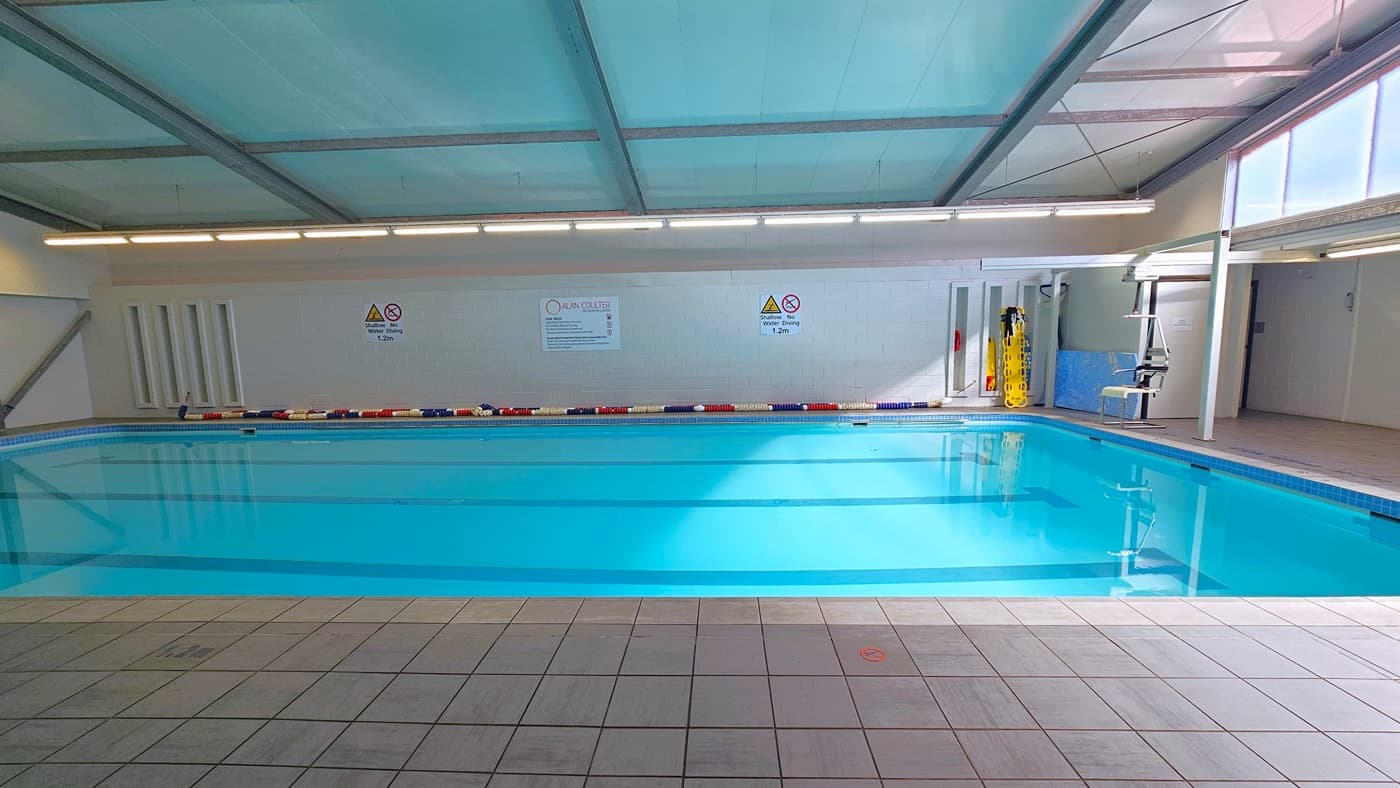The 16m swimming pool has colourful floating ropes on the pool deck. Banners and safety reminders are affixed to the middle wall. Hydrotherapy equipment and a hoist are positioned on the right. The low ceiling is white with exposed steel beams, lined with fluorescent lights. The floor is made of white tiles.