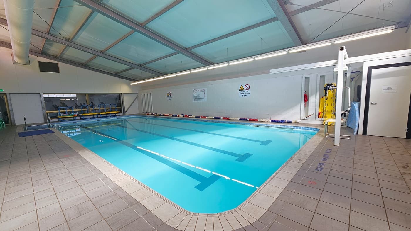 The 16m swimming pool has colourful floating ropes on the pool deck. Banners and safety reminders are affixed to the right wall. An entrance to the pool through stairs with a railing and a ramp is positioned in the left corner. Hydrotherapy equipment and a hoist are placed on the right. The sloping ceiling is white with exposed steel beams, large air conditioning vents, and lined with fluorescent lights. The floor is made of white tiles.