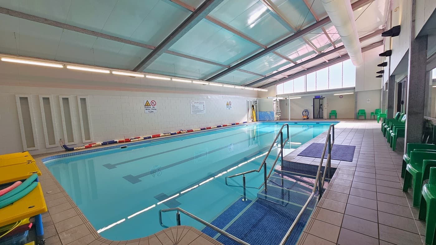 The 16m swimming pool has colorful floating ropes on the pool deck. Banners and safety reminders are affixed to the left wall. An entrance to the pool through stairs with a railing and a ramp is positioned in the corner. Hydrotherapy equipment and a hoist are visible at the far end. Plastic tables hold swimming accessories on the left, while plastic green chairs are aligned to the right. The ceiling is white with exposed steel beams, and large air conditioning vents.