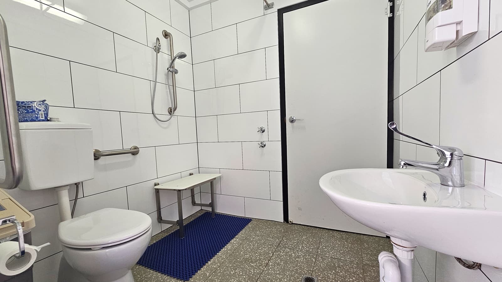 Inside the accessible bathrooms, the toilet bowl is positioned on the left, equipped with handrails. A tissue dispenser and a rubbish bin are beside it. Adjacent is the shower with a bench and a blue anti-slippery mat. In the middle is the white manual door. To the right, there is a washbasin and a liquid soap dispenser mounted to the wall. The floor has grey granite design tiles, and the entire wall has white tiles.