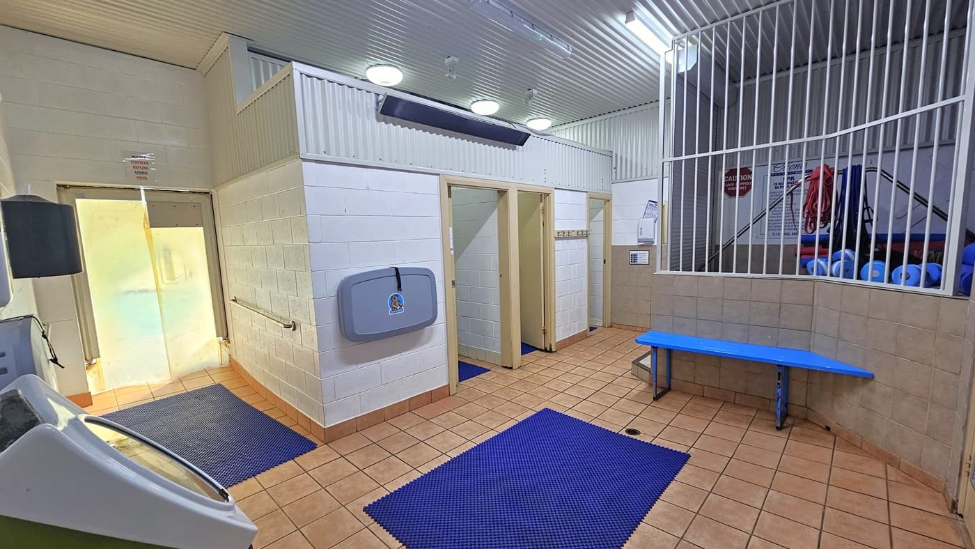 To the right are two shower rooms. A foldable baby change table is in the middle. There is a blue anti-slip mat on the left. Faulty exercise machines are lined up on the left side. On the right, a storage for gym equipment is enclosed by metal grills. A blue bench chair is placed below it. The floor is light pink tiles, and the wall is white with a corrugated design.