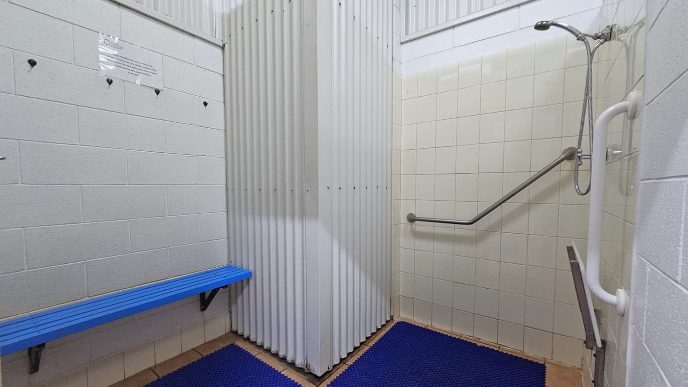A closer look inside the shower room reveals the shower mounted on the right wall, accompanied by handrails. A foldable bench is positioned underneath. To the left, a blue bench chair is affixed to the wall. The floor is covered with a blue anti-slippery mat. The wall is a combination of white tiles.