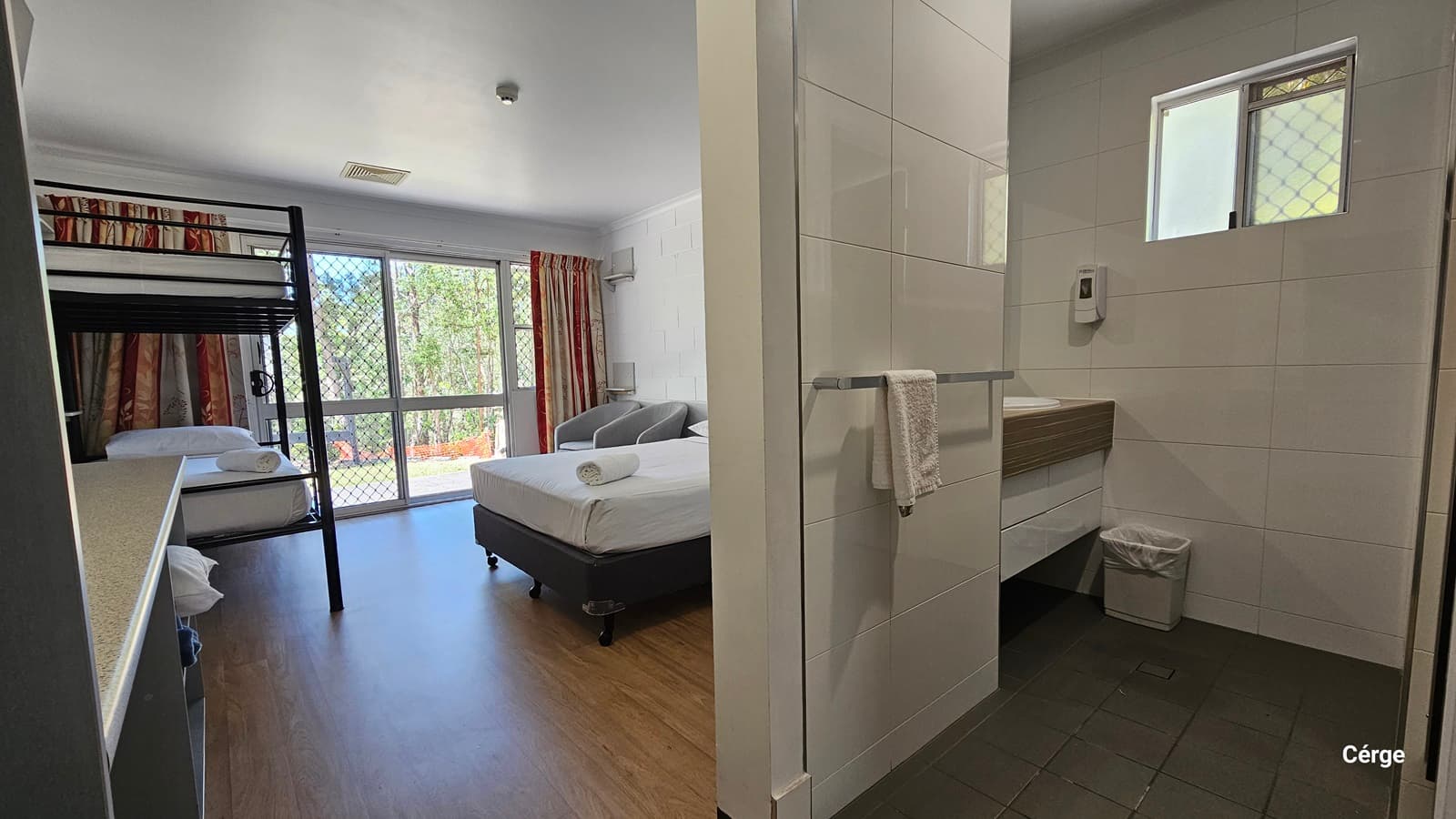 Inside the room, to the left is a double bed with white linens. Beside it is a cushioned grey chair positioned near the glass window. To the right is a double-deck bed. Adjacent to it is a table. The ceiling and walls are all white with a brick design. The floor has a timber design. To the right is the ensuite toilet featuring grey tiles and white walls. A washbasin and rubbish bin are positioned in the corner.