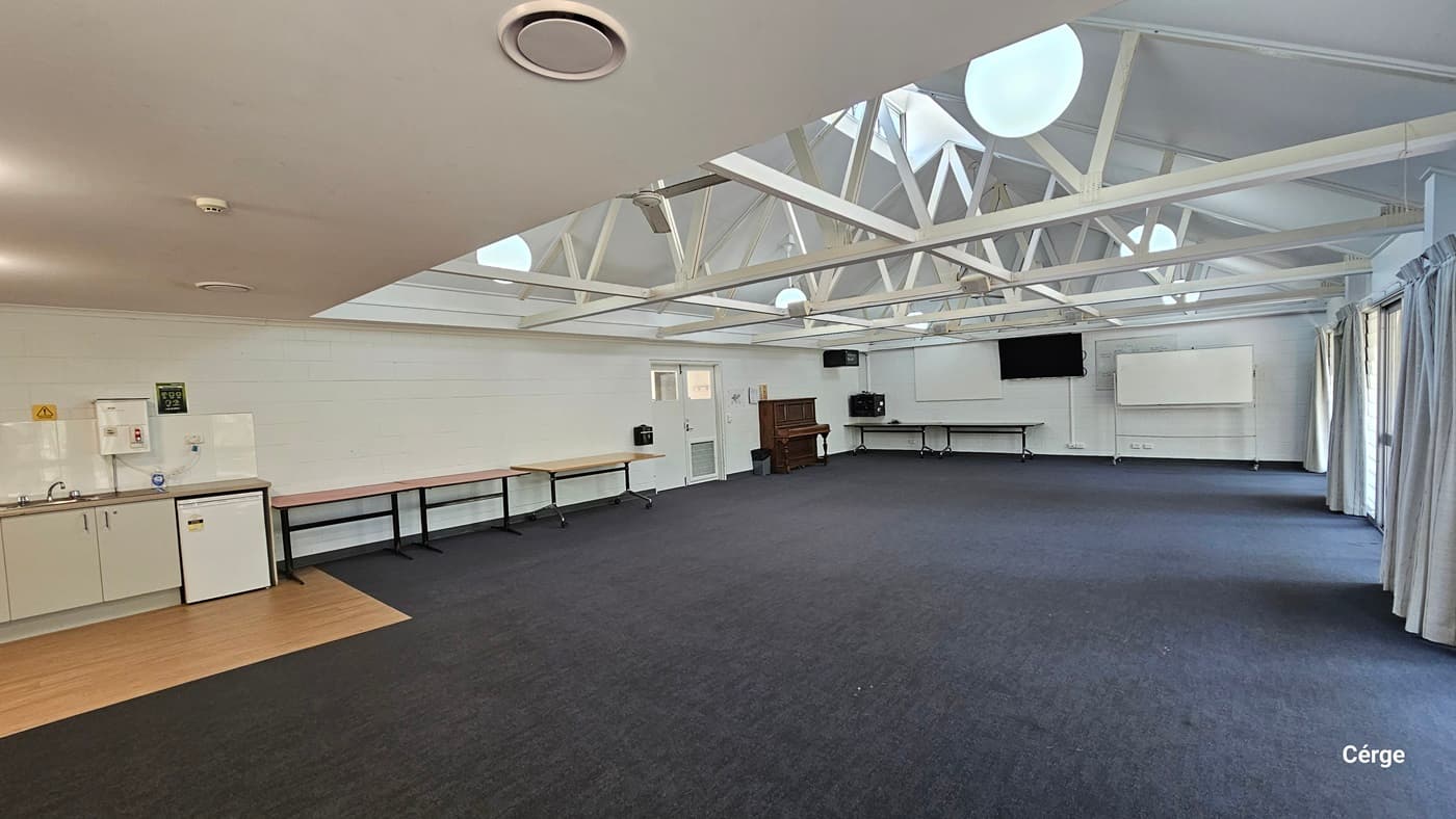 Inside the meeting room, a drinking fountain is situated to the left. A small section of timber tiles is installed on the floor. Tables are arranged against the wall. At the far end, there is a wooden piano, a flatscreen TV, and a movable whiteboard. To the right, floor-to-ceiling curtains cover the glass wall. The ceiling features an industrial look with exposed white metal beams and pendant lights. The floor has dark grey tiles.