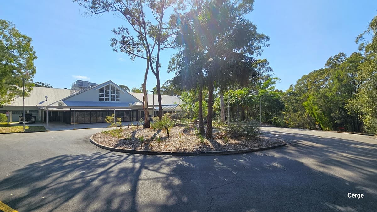 A paved area with a circular garden bed in the centre, surrounded by a road. Sparse trees and shrubbery populate the garden bed and the periphery. Behind is a building with a large triangular roof feature and an extended wing. Tall green trees frame the background.
