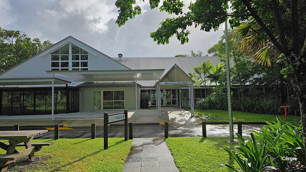 A building with a gabled roof and large windows on the left, featuring a covered entrance with a sign reading "Welcome to the Park" in the middle. The right side has lush greenery, including trees and bushes. In the foreground, there is a picnic table on a grassy area and a paved walkway leading to the entrance. The background includes more trees and foliage. The floor is a mix of grass and pavement.
