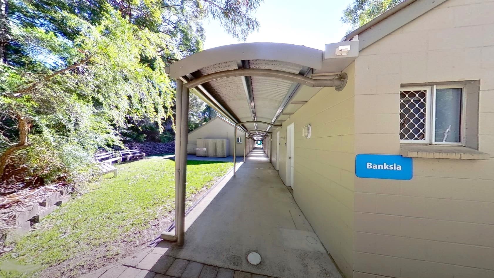 The Banksia accommodation is a low-rise cream building with a small window and an extended roofing. To the left, trees and grass surround the area. Bench seatings are arranged under the shades of huge trees.
