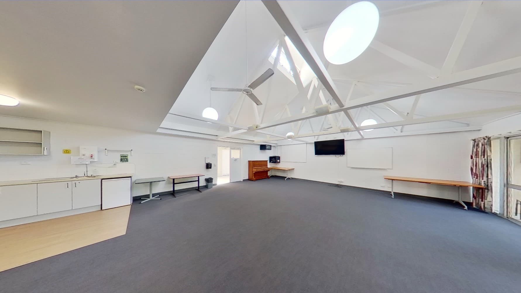 Inside the meeting room, a drinking fountain is positioned on the left. Tables line the left wall, with a flat-screen TV and a whiteboard mounted on the middle wall. A wooden piano sits in the corner. At the far end is a floor-to-ceiling glass window with curtains. Additional tables are against the right wall. The ceiling boasts an industrial look with exposed white metal beams and pendant lights. Dark grey tiles cover the floor.